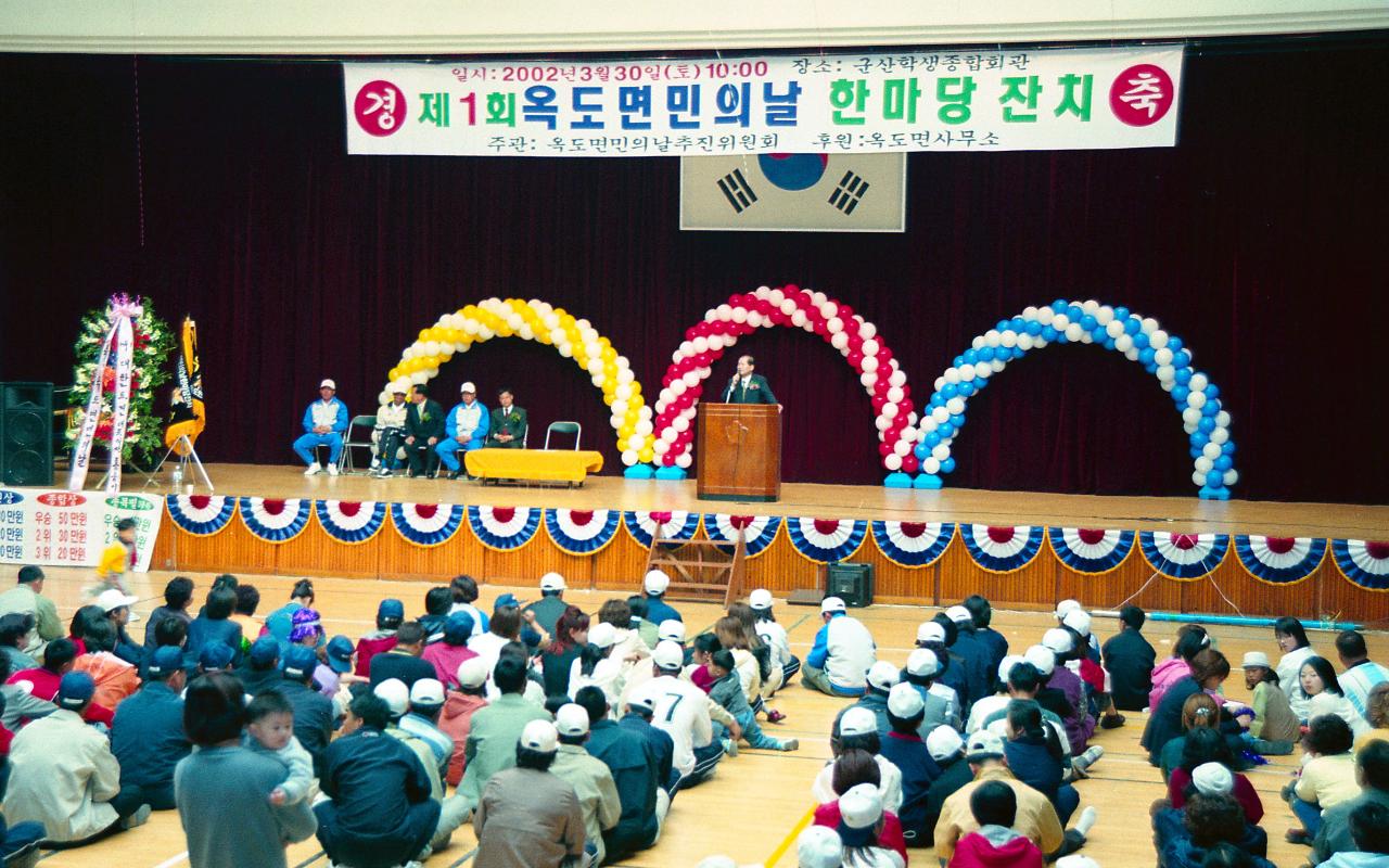 옥도면민 체육대회가 열리는 체육관의 무대 위에서 말씀을 하고 계신 시장님과 바닥에 앉아 듣고 있는 면민들