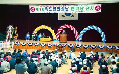 옥도면민 체육대회가 열리는 체육관의 무대 위에서 말씀을 하고 계신 시장님과 바닥에 앉아 듣고 있는 면민들