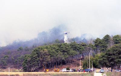 불이 붙은 곳에 물을 뿌리고 있는 헬리콥터와 산 아래 모여있는 사람들과 차들