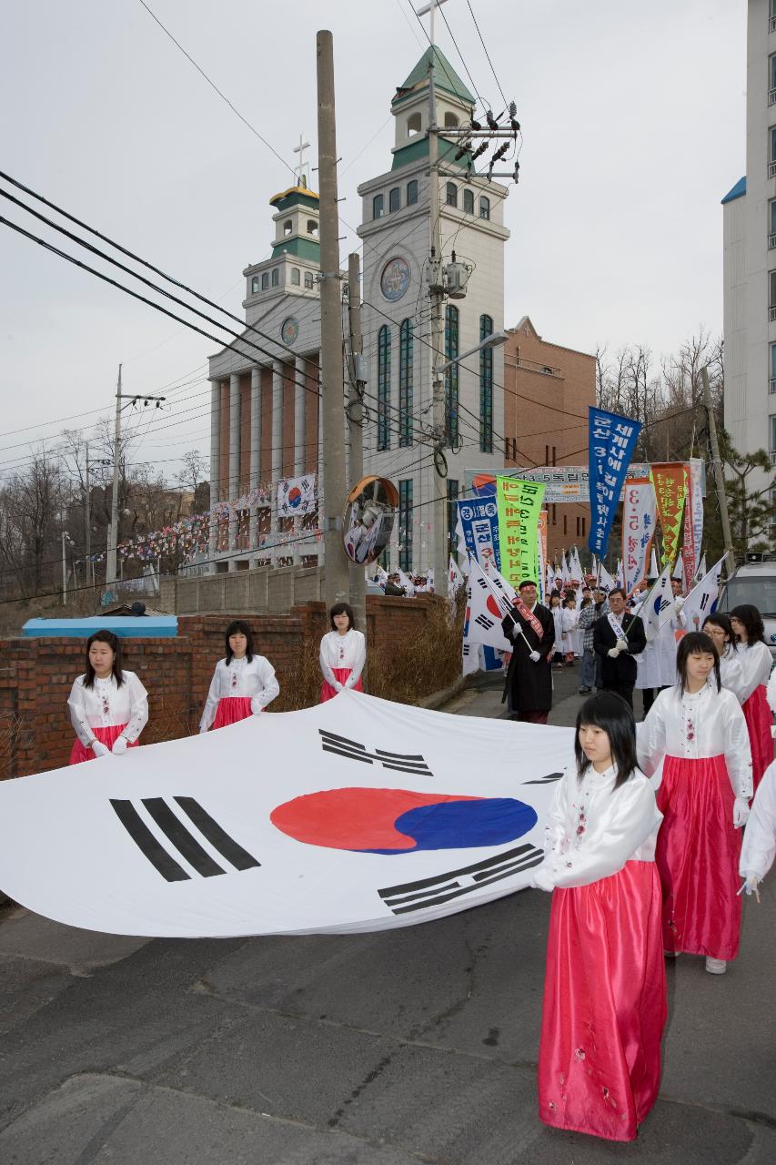 큰 태극기들을 들고 행진하는 빨간색 치마 한복을 입은 참가자들과 뒤로 따라오는 하얀 옷을 입고 태극기를 들고있는 시민들