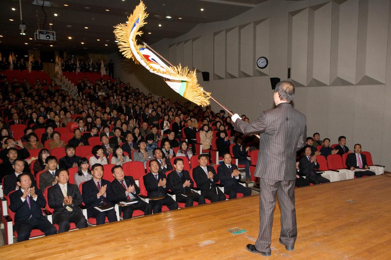 무대위에서 깃발을 흔들고 있는 관계자와 자리에 앉아 박수를 치고 있는 관련인사들